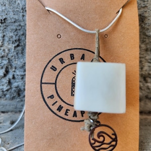 May include: A silver snake chain necklace featuring a square white stone pendant suspended by a tan cord. A small circular charm with a wave design hangs below the stone. The necklace is displayed on a brown card printed with the text URBAN PINEAPPLE.