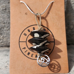May include: A necklace featuring three stacked, circular, black and white patterned beads suspended from a silver chain. A small, circular silver charm with a cut-out wave design hangs at the bottom. The necklace is displayed on a tan card with the text URBAN PINEAPPLE.