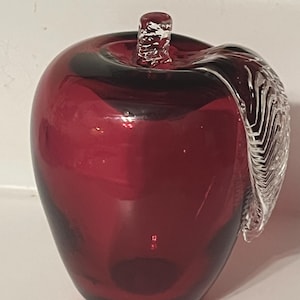 Może przedstawiać: Szklana figurka jabłka w kolorze głębokiej czerwieni o gładkim, półprzezroczystym korpusie. Na górze znajduje się mała, teksturowana łodyga z przezroczystego szkła oraz wyraźny liść z przezroczystego szkła z detalami białych linii.