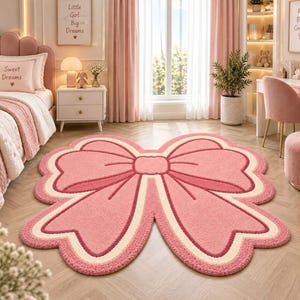 Puede incluir: Una alfombra grande con forma de lazo que presenta un centro rosa suave, un borde color crema y un contorno rosa más oscuro. La alfombra tiene una superficie texturizada y afelpada, con cuatro bucles redondeados que forman un lazo decorativo.