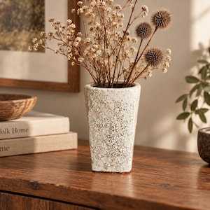 Pode incluir: Um vaso de cerâmica alto e cónico com um acabamento texturizado branco e creme rachado. O vaso tem uma base quadrada e contém um ramo de flores secas, gipsofila castanho-clara e cabeças de sementes redondas e espinhosas em tons naturais.