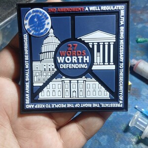 Può includere: Una toppa quadrata in gomma blu scuro e bianca con un grafico del Campidoglio e della Casa Bianca. Il testo recita: 2ND AMENDMENT: A WELL REGULATED MILITIA, BEING NECESSARY TO THE SECURITY OF A FREE STATE, THE RIGHT OF THE PEOPLE TO KEEP AND BEAR ARMS, SHALL NOT BE INFRINGED. 27 WORDS WORTH DEFENDING.