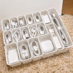 Op de afbeelding: Een witte kunststof lade-organizer met meerdere rechthoekige vakken met diverse witte elektronische accessoires, waaronder oplaadkabels, stroomadapters, bedrade oortjes en een oplaadcase voor draadloze oortjes.