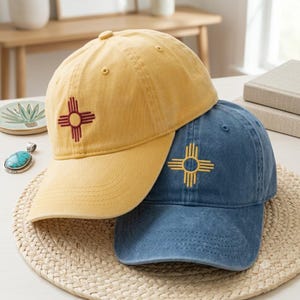 Può includere: Un cappellino da baseball giallo con un simbolo solare Zia ricamato in rosso e un cappellino da baseball blu con un simbolo solare Zia ricamato in giallo. Entrambi i cappellini presentano una struttura a sei pannelli con visiera curva.