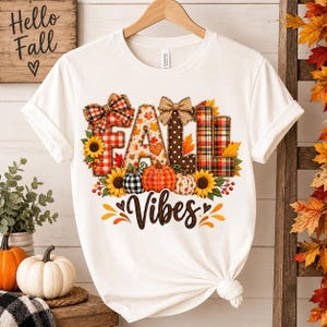 May include: A white short-sleeve t-shirt featuring a graphic print of the word FALL in large, patterned block letters decorated with plaid, polka dots, and autumn leaves. Below, the word Vibes is written in brown script with pumpkins.