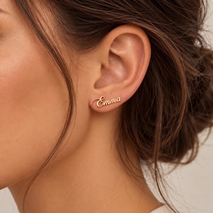 May include: A gold-toned stud earring featuring the name Emma written in a cursive script font. The metal surface has a polished finish, and the name is oriented horizontally along the earlobe, secured with a standard post backing.