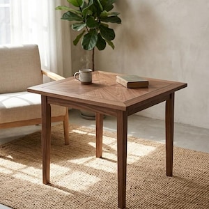 Puede incluir: Una mesa cuadrada de madera con un acabado marrón intenso y un patrón de veta distintivo en la superficie. La mesa cuenta con cuatro patas rectas y una superficie plana, que sostiene una taza de cerámica y un libro cerrado de tapa clara.