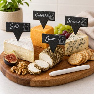 May include: Four black slate cheese markers with white text reading Brie, Cheddar, Goat, and Stilton, each featuring a small mouse illustration. The markers are inserted into wedges of Brie, orange Cheddar, herb-crusted goat cheese, and blue Stilton.