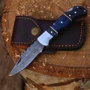 May include: A folding pocket knife with a Damascus steel blade featuring a wavy pattern, a blue wood handle with white dot inlays, and brass accents. The knife is paired with a dark brown leather sheath featuring yellow stitching.