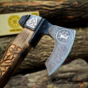 Könnte beinhalten: Ein Handbeil mit einem dunklen Metallkopf, der ein silbernes Valknut-Symbol, eine kreisförmige Gravur eines Reiters und komplizierte Knotenmuster aufweist. Der Holzgriff ist mit Blumenmustern verziert und hat ein dunkles, verwittertes Finish.