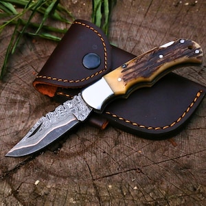 Op de afbeelding: Een inklapbaar zakmes met een patroon van Damascusstaal en een gestructureerd bruin gewei-handvat, voorzien van een zilveren bolster en drie metalen pinnen, vergezeld door een donkerbruine lederen schede met oranje stiksels.