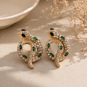 May include: A pair of gold-toned snake-shaped earrings featuring clear pavé rhinestones and marquise-cut emerald green stones. Each earring has a black stone for the eye and is designed in a coiled, circular position with a textured metal finish.