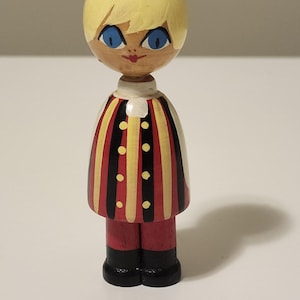 Puede incluir: Una muñeca de madera con cabeza redondeada, cabello rubio pintado y ojos azul brillante. La figura viste una túnica con rayas verticales rojas, amarillas y negras con pequeños puntos amarillos en el centro, pantalones rojos y zapatos negros.