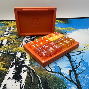 May include: A rectangular orange plastic storage box with an open lid, containing a collection of translucent orange and yellow six-sided dice. The dice feature white numerical markings and mathematical symbols, arranged in a grid within the box.