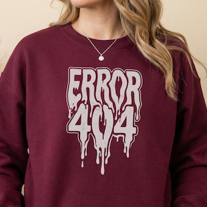 May include: A maroon long-sleeve crewneck sweatshirt featuring a large, light gray graphic on the chest. The graphic displays the text "ERROR 404" in a stylized, dripping font that appears to be melting downward, centered on the front of the garment.