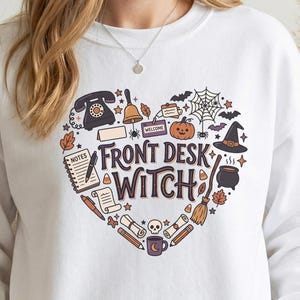 May include: A white crewneck sweatshirt featuring a heart-shaped graphic containing the text FRONT DESK WITCH. The design includes illustrations of a rotary phone, bell, pumpkin, spider web, witch hat, cauldron, broom, skull, and office supplies.