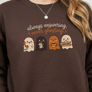 May include: A dark brown crewneck sweatshirt featuring a graphic of four cartoon ghosts. The text above reads always answering, never ghosting. The ghosts are depicted holding a telephone, a clipboard, a coffee mug, and a welcome sign respectively.