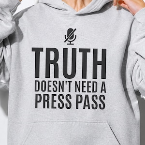 May include: A light gray hooded sweatshirt featuring a black graphic of a microphone with a slash through it, positioned above the text TRUTH DOESN'T NEED A PRESS PASS in bold, black, sans-serif capital letters centered on the front chest area.