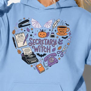 May include: A light blue hooded sweatshirt featuring a heart-shaped graphic with the text "SECRETARY WITCH". The design includes a typewriter, a rotary phone, a to-do list, a stapler, a pumpkin, a coffee cup, scissors, and various office supplies.