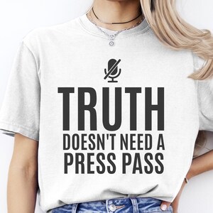 May include: A white short-sleeved t-shirt featuring a black graphic of a microphone with a slash through it. Below the icon, the text reads TRUTH DOESN'T NEED A PRESS PASS in bold, black, sans-serif capital letters centered on the chest area.