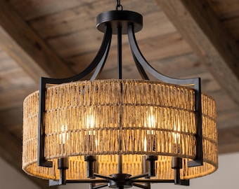 May include: A drum-shaped chandelier featuring a matte black metal frame and a cylindrical shade wrapped in natural tan jute rope. The fixture includes six vertical light sockets arranged in a circle, suspended by a black chain.