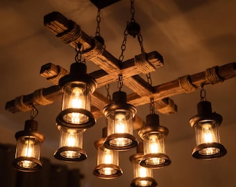 May include: A ceiling chandelier featuring a cross-shaped wooden beam structure wrapped with rope, suspended by metal chains. Seven lantern-style light fixtures with clear glass cylinders and black metal frames hang from the beams, each containing a glowing bulb.