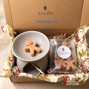 Op de afbeelding: Een bruine kartonnen geschenkdoos met een matte beige keramische waxinelichthouder, een lichtbruin wassmeltblokje in de vorm van een peperkoekmannetje en een doorzichtig plastic zakje met extra wassmeltblokjes voorzien van het Kindle-logo.