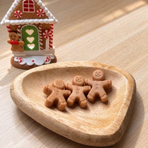Può includere: Una piccola ciotola in legno marrone chiaro contenente tre cera profumata a forma di omino di pan di zenzero. Sullo sfondo, una statuetta decorativa a forma di casetta di pan di zenzero presenta una porta verde, glassa bianca e decorazioni.