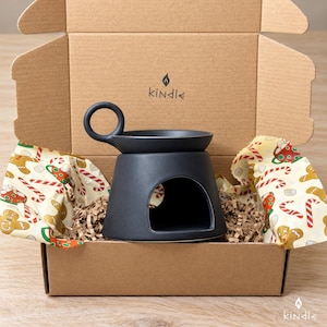Op de afbeelding: Een matzwarte keramische waxwarmer met een cirkelvormig handvat en een brede opening aan de basis, geplaatst in een bruine kartonnen doos. De doos bevat de tekst "kindle" met een vlamicoon en is bekleed met papier met peperkoekpatroon.