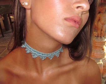Könnte beinhalten: Eine hellblaue Häkel-Halskette als Choker mit einem zarten, bogenförmigen Randdesign. Die Halskette ist aus feinem Garn gefertigt und liegt eng am Hals an, wobei sie ein detailliertes Webmuster mit kleinen, sich wiederholenden Schlaufen zeigt.