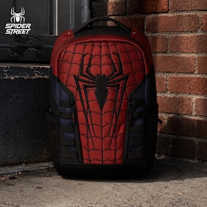 May include: A red and blue backpack featuring a black spider emblem and a web pattern on the front panel. The bag includes black side panels, vertical black webbing straps, and a top handle. The text SPIDER STREET is visible in the top left corner.