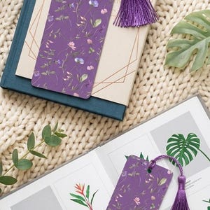 May include: Two rectangular purple bookmarks featuring a delicate floral pattern of small pink, blue, and green botanical illustrations. Each bookmark includes a hole at the top with a matching purple silk tassel attached to the end.