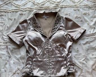 Può includere: Una camicetta in raso color champagne con colletto dentellato decorato con borchie metalliche color argento. Il capo presenta maniche corte, una chiusura centrale anteriore e dettagli decorativi con lacci in stile corsetto sui fianchi.