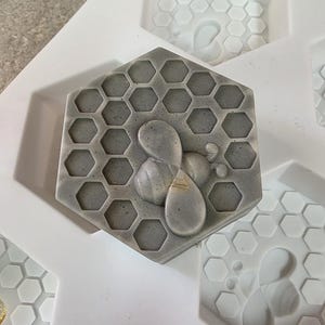 May include: A hexagonal gray soap bar featuring a raised bee design and a honeycomb pattern on its surface, resting inside a white silicone mold with multiple empty hexagonal cavities that also feature bee and honeycomb impressions.