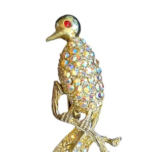Könnte beinhalten: Goldfarbene Vogelbrosche mit schillernden Strasssteinen. Der Vogel hat ein rotes Auge und sitzt auf einem Ast. Die Brosche ist ein Vintage-Stück, vermutlich aus der Mitte des 20. Jahrhunderts.