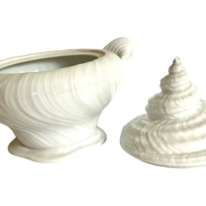 Pu&ograve; includere: Scodella da portata in ceramica bianca a forma di conchiglia con coperchio abbinato. La ciotola presenta un design testurizzato a spirale e un manico su un lato.
