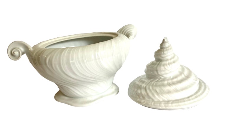 Pu&ograve; includere: Scodella da portata in ceramica bianca a forma di conchiglia con coperchio abbinato. La ciotola presenta un design testurizzato a spirale e un manico su un lato.