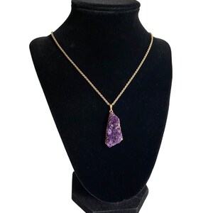 May include: A gold chain necklace with a large, purple amethyst pendant. The pendant is a cluster of crystals in a rough, natural shape.