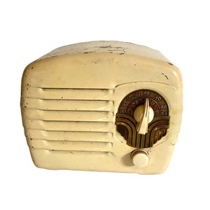 Könnte beinhalten: Cremefarbenes Vintage-Radio mit abgerundeter Oberseite und horizontalen, schlitzartigen Linien. Das Radio verfügt über ein Zifferblatt mit Zahlen und einen Drehknopf. Das Zifferblatt hat ein goldfarbenes Dekor. Die Oberfläche des Radios weist Gebrauchsspuren auf.