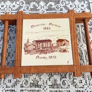 Könnte beinhalten: Ein holzgerahmtes Kachel mit einer Sepia-Illustration von Gebäuden und dem Text "Norsebo, Norwegen 1882" und "Norsby, 1978". Der Rahmen ist aus hellbraunem Holz und hat einen Ständer.