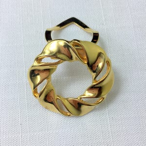 Könnte beinhalten: Goldfarbene Metallbrosche mit einem gedrehten, kreisförmigen Design. Die Brosche hat einen schlichten, eleganten Stil.