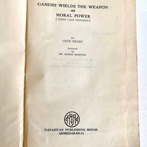 May include: Open book with the title "GANDHI WIELDS THE WEAPON OF MORAL POWER" by Gene Sharp. The foreword is by Dr. Albert Einstein. The book is published by Navajivan Publishing House, Ahmedabad-14.