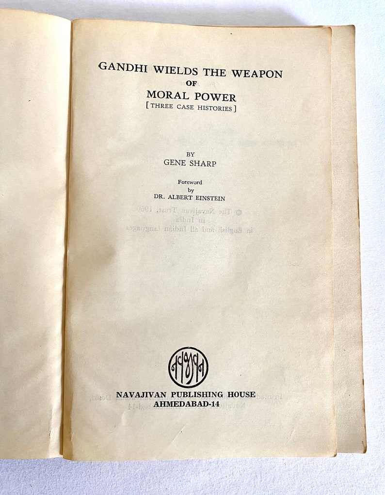 May include: Open book with the title "GANDHI WIELDS THE WEAPON OF MORAL POWER" by Gene Sharp. The foreword is by Dr. Albert Einstein. The book is published by Navajivan Publishing House, Ahmedabad-14.