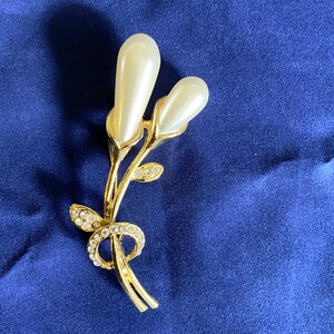 Könnte beinhalten: Eine goldfarbene Brosche mit zwei weißen, perlmuttartigen Blütenknospen und einem Stiel mit klaren Strasssteinen.