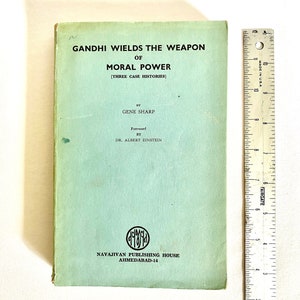 May include: A vintage book titled "GANDHI WIELDS THE WEAPON OF MORAL POWER" with text by Gene Sharp and a foreword by Dr. Albert Einstein. The book has a light green cover with black text and a logo. A ruler is placed next to the book.
