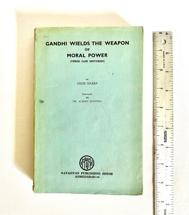 May include: A vintage book titled "GANDHI WIELDS THE WEAPON OF MORAL POWER" with text by Gene Sharp and a foreword by Dr. Albert Einstein. The book has a light green cover with black text and a logo. A ruler is placed next to the book.