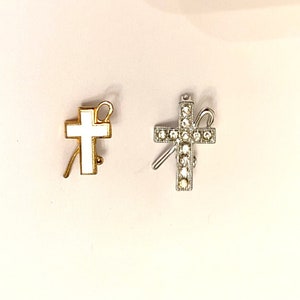 Könnte beinhalten: Zwei silberne und goldene Kreuznadeln mit einem weißen Emaille-Kreuz und einem Hirtenstab-Design. Das silberne Kreuz ist mit Strasssteinen verziert.