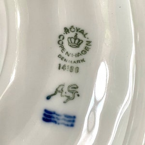 Pu&ograve; includere: Primo piano di un marchio di porcellana Royal Copenhagen con le parole "Royal Copenhagen Denmark" e il numero "14180" in verde. Il marchio include anche una corona e le lettere "A/S" in blu.