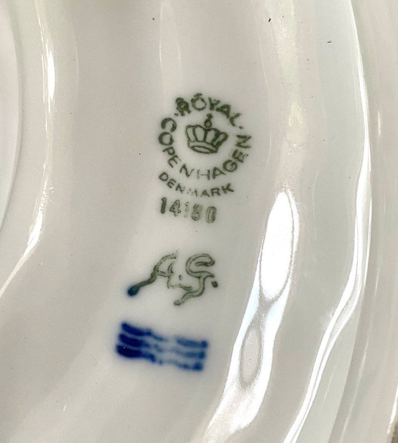 Pu&ograve; includere: Primo piano di un marchio di porcellana Royal Copenhagen con le parole "Royal Copenhagen Denmark" e il numero "14180" in verde. Il marchio include anche una corona e le lettere "A/S" in blu.
