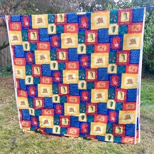 Könnte beinhalten: Ein farbenfroher Stoff mit einem sich wiederholenden Muster aus gelben, blauen, roten und grünen Quadraten. Jedes Quadrat enthält ein weihnachtliches Bild, wie eine Tannenzapfen, einen Stern oder einen Stechpalmenzweig. Der Stoff hängt draußen.